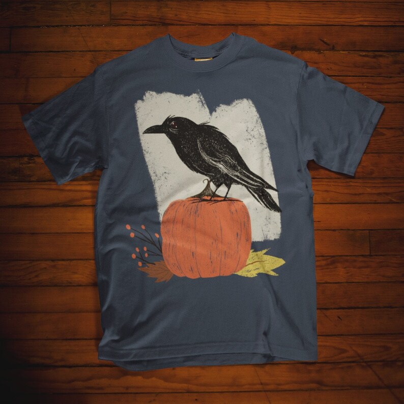 May include: A dark blue t-shirt with a graphic of a black crow perched on top of an orange pumpkin. The graphic is surrounded by fall foliage.