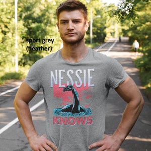 May include: A heather grey t-shirt with a graphic of Nessie, the Loch Ness Monster, and the text "Nessie Knows".