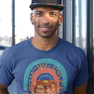 Puede incluir: Un hombre con una camiseta azul real con un gr&aacute;fico de arco&iacute;ris de estilo vintage y el texto "The Love is Real" debajo de una pila de panqueques con jarabe y mantequilla. Lleva una gorra de b&eacute;isbol negra con una franja roja, blanca y azul.