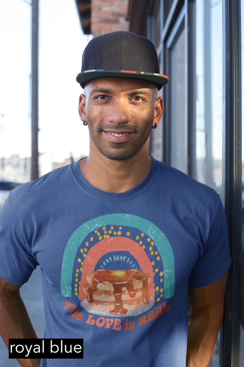 Puede incluir: Un hombre con una camiseta azul real con un gr&aacute;fico de arco&iacute;ris de estilo vintage y el texto "The Love is Real" debajo de una pila de panqueques con jarabe y mantequilla. Lleva una gorra de b&eacute;isbol negra con una franja roja, blanca y azul.