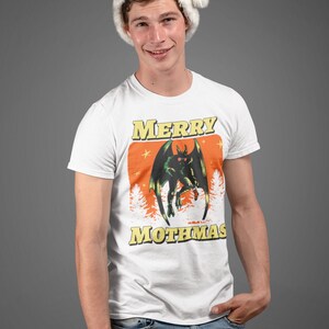 May include: A white t-shirt with a green monster graphic and the text "Merry Mothmas". The person wearing the shirt is also wearing a red Santa hat.