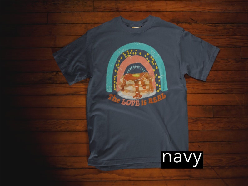 Puede incluir: Camiseta azul marino con un gr&aacute;fico de arco&iacute;ris y una pila de panqueques con jarabe. El texto "The Love is Real" est&aacute; impreso en la camiseta.