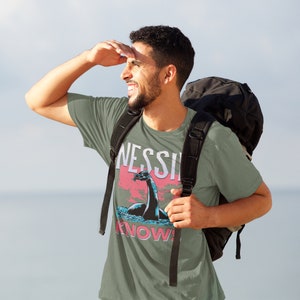 May include: A green t-shirt with a graphic of a Loch Ness Monster and the text "NESSIE KNOWS!" printed on it. The shirt is being worn by a person with a black backpack.