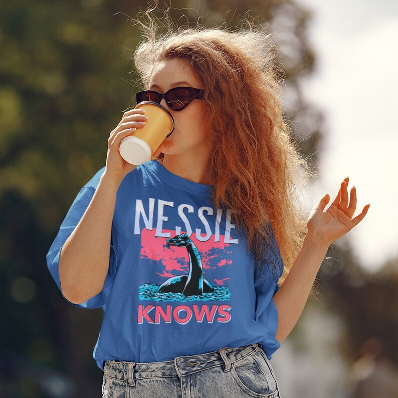 May include: A woman wearing a blue t-shirt with a graphic of a Loch Ness Monster and the text "Nessie Knows".