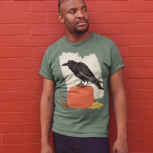 May include: A green t-shirt with a vintage-style illustration of a black raven perched on a pumpkin. The illustration is surrounded by fall-themed foliage.