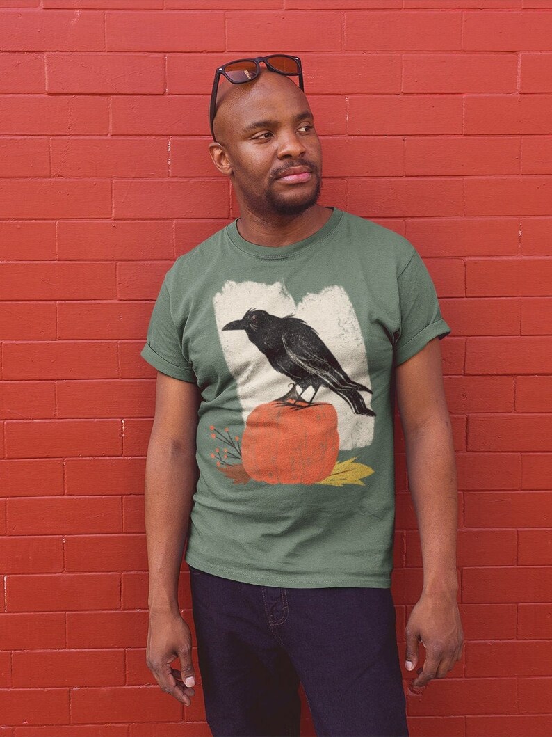 May include: A green t-shirt with a vintage-style illustration of a black raven perched on a pumpkin. The illustration is surrounded by fall-themed foliage.
