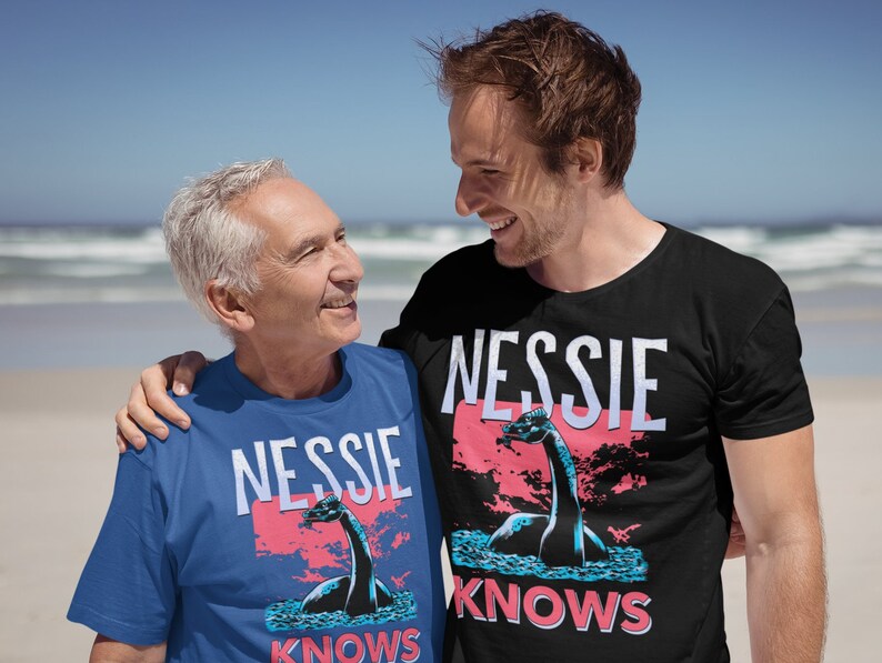 May include: Two people wearing blue and black t-shirts with a graphic of a Loch Ness Monster and the text "Nessie Knows".
