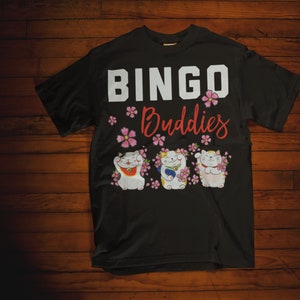 May include: Black t-shirt with the text "Bingo Buddies" in white and red lettering. Three cartoon lucky cats with pink flowers are below the text.