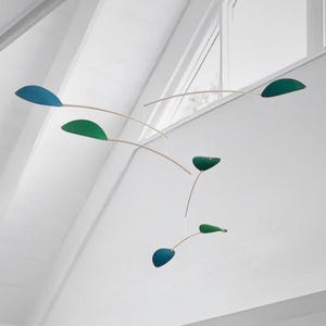 Puede incluir: Una escultura móvil con piezas en forma de hoja de color verde azulado y verde suspendidas de finos cables. La móvil está colgada del techo de una habitación con paredes blancas y una gran ventana.