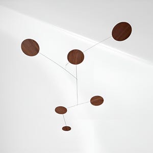 May include: A modern mobile with six dark brown, circular wooden elements suspended from thin, silver-colored wires. The mobile is designed to hang from the ceiling, creating a minimalist and artistic decorative piece. The wooden circles vary in size.
