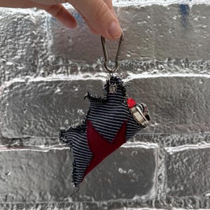 Puede incluir: Un llavero hecho a mano con un diseño único. El llavero tiene una tela a rayas azul oscuro y blanco con un acento en forma de estrella roja. Está unido a un mosquetón plateado.