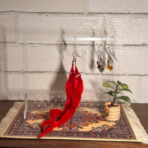 May include: A clear acrylic earring stand with three hooks holding various earrings. A red fuzzy earring dangles from one hook. A small potted plant sits on a patterned rug in front of the stand.