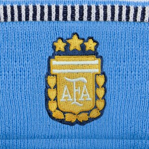May include: Close-up of a light blue knitted fabric with a navy and white striped band at the top. A gold, navy, and white embroidered emblem featuring three stars and the letters "AFA" is centered on the fabric.