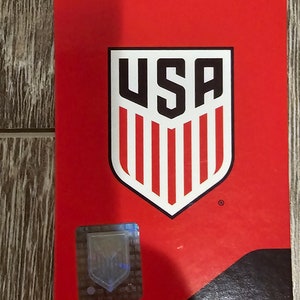 May include: Red tag with a white and black USA shield logo. The shield has red and white stripes and the letters USA in black. The tag has a black border and a small holographic sticker.