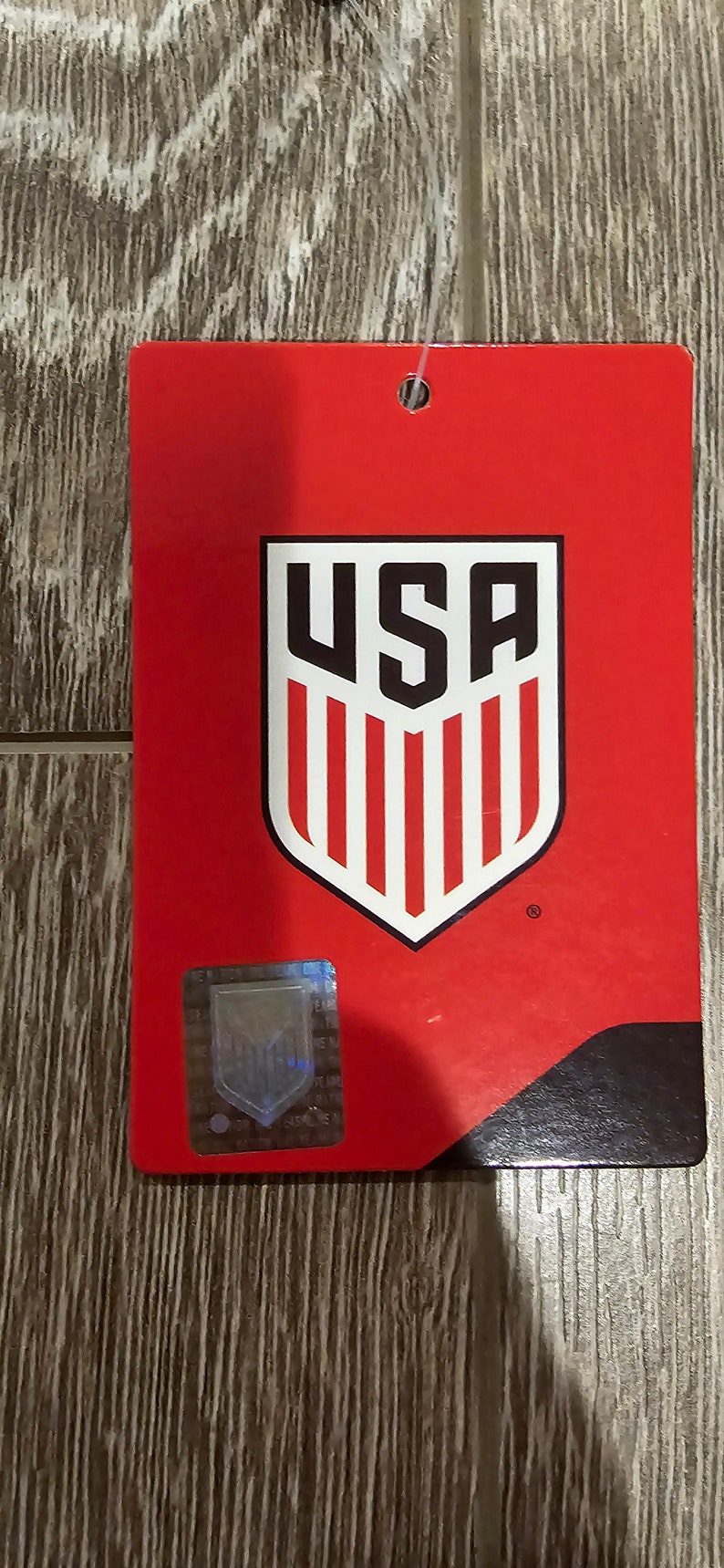 May include: Red tag with a white and black USA shield logo. The shield has red and white stripes and the letters USA in black. The tag has a black border and a small holographic sticker.