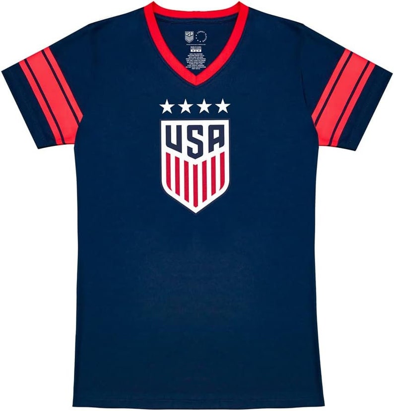May include: A navy blue t-shirt with red and white stripes on the sleeves. The shirt has a v-neck and features a white shield with the word "USA" and five stars above it.