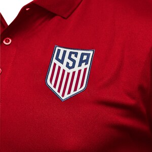 May include: Red polo shirt with a white and blue embroidered patch featuring the word "USA" and red and white stripes.