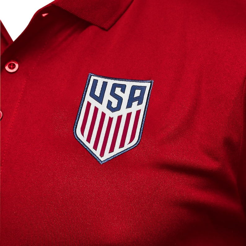 May include: Red polo shirt with a white and blue embroidered patch featuring the word "USA" and red and white stripes.