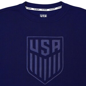 Op de afbeelding: Een marineblauw sweatshirt met een groot, ge&euml;mbost, grijs schildvormig logo met het woord "USA" en verticale strepen. De tekst "U.S. SOCCER" wordt herhaald rond de halslijn.