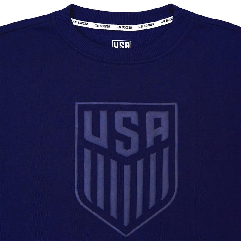 Op de afbeelding: Een marineblauw sweatshirt met een groot, ge&euml;mbost, grijs schildvormig logo met het woord "USA" en verticale strepen. De tekst "U.S. SOCCER" wordt herhaald rond de halslijn.