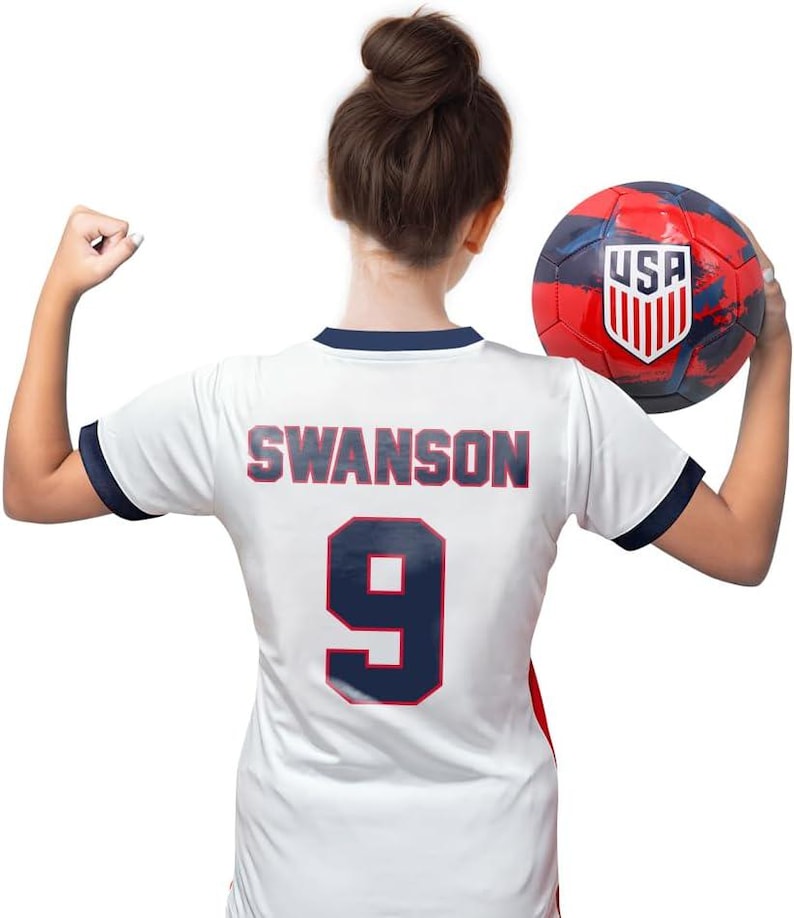 Puede incluir: Camiseta de f&uacute;tbol blanca con ribetes azules y el nombre "SWANSON" y el n&uacute;mero "9" impresos en la espalda. La camiseta la sostiene una persona que sostiene un bal&oacute;n de f&uacute;tbol rojo, blanco y azul con la palabra "USA" impresa en &eacute;l.