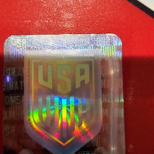 May include: A holographic sticker with a shield-shaped design featuring the word "USA" in gold lettering. The shield is surrounded by a rainbow-colored border.