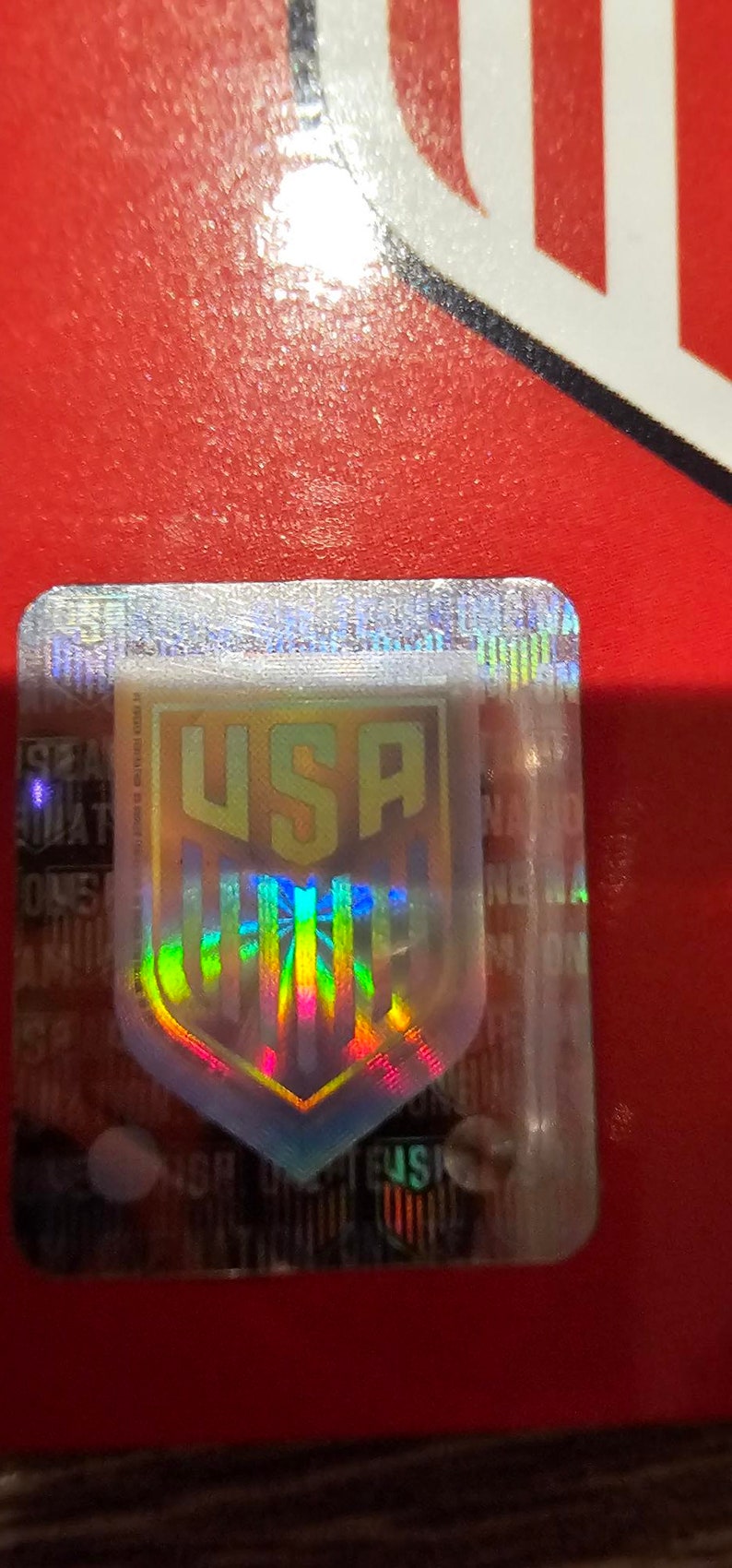 May include: A holographic sticker with a shield-shaped design featuring the word "USA" in gold lettering. The shield is surrounded by a rainbow-colored border.