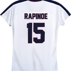 K&ouml;nnte beinhalten: Wei&szlig;es Fu&szlig;balltrikot mit marineblauem und rotem Besatz. Auf der R&uuml;ckseite des Trikots sind der Name "RAPINOE" und die Nummer "15" in Marineblau mit roten Umrandungen gedruckt.
