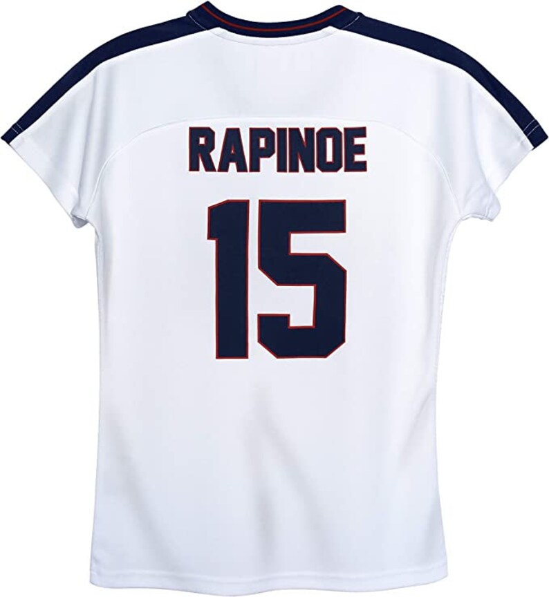 K&ouml;nnte beinhalten: Wei&szlig;es Fu&szlig;balltrikot mit marineblauem und rotem Besatz. Auf der R&uuml;ckseite des Trikots sind der Name "RAPINOE" und die Nummer "15" in Marineblau mit roten Umrandungen gedruckt.
