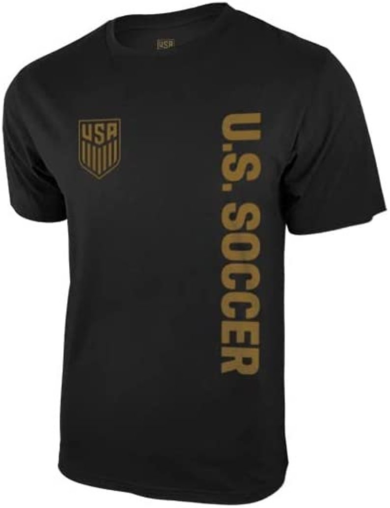 以下が含まれることがあります： 右側に金色のU.S. Soccerのロゴとテキストが書かれた黒いTシャツ。ロゴは、中にUSAの文字が書かれた盾です。