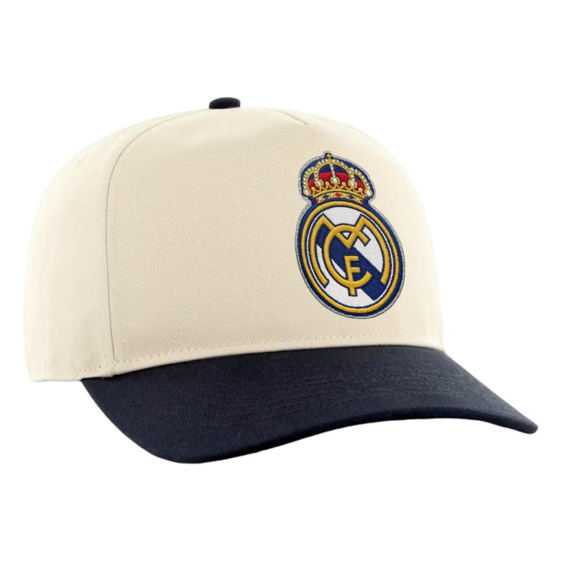 May include: A cream-colored baseball cap with a navy blue brim. The cap features an embroidered emblem with a crown and the letters "RMCF" in a circular design, showcasing a team logo.