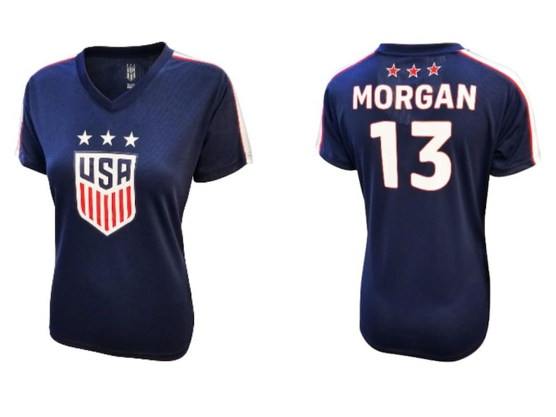 K&ouml;nnte beinhalten: Ein marineblaues Fu&szlig;balltrikot mit wei&szlig;en und roten Akzenten. Das Trikot hat drei Sterne und "USA" auf der Vorderseite und "MORGAN 13" auf der R&uuml;ckseite.
