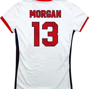 May include: White soccer jersey with red and blue trim. The back of the jersey has the name "MORGAN" and the number "13" in red with a blue outline.