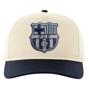 May include: Cream-colored baseball cap with a navy blue brim and a black button on top. The cap features an embroidered crest with the letters "FCB" and a soccer ball design in navy blue and light blue.