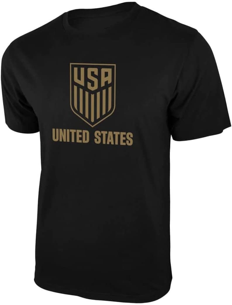 May include: Black t-shirt featuring a gold United States soccer team logo. The logo depicts the letters "USA" within a shield with vertical stripes.