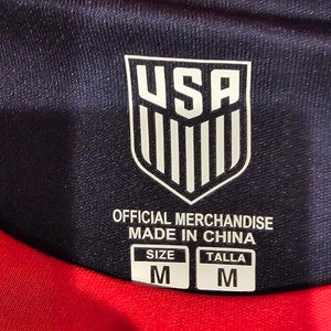 May include: A black and red soccer jersey with a white USA logo and the text "Official Merchandise Made in China". The jersey has a size tag that says "Size M Talla M".