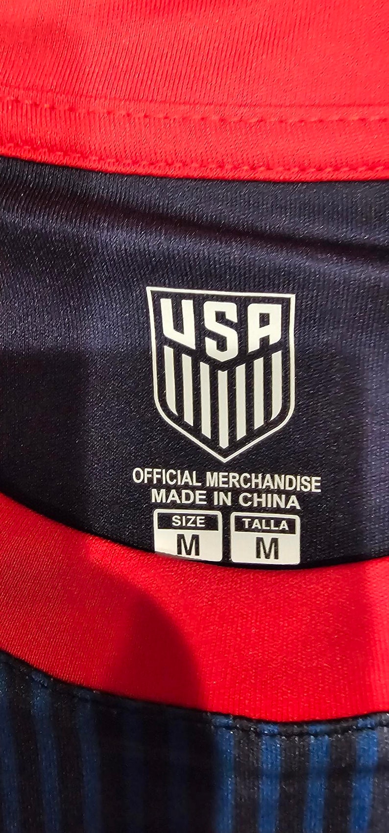 May include: A black and red soccer jersey with a white USA logo and the text "Official Merchandise Made in China". The jersey has a size tag that says "Size M Talla M".