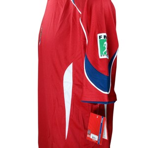 May include: Red soccer jersey with white and blue accents. The jersey has a small logo on the left chest and a larger logo on the right sleeve that says "FIFA Official".