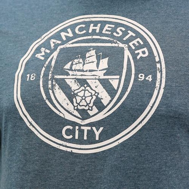 May include: A white distressed vintage logo on a blue background. The logo says "Manchester City" with a ship and a rose inside a shield. The numbers "18" and "94" are on either side of the shield.