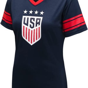 May include: A navy blue short-sleeved t-shirt with red and white accents. The shirt features a white shield with the word "USA" and five stars above it. The sleeves have two red stripes.