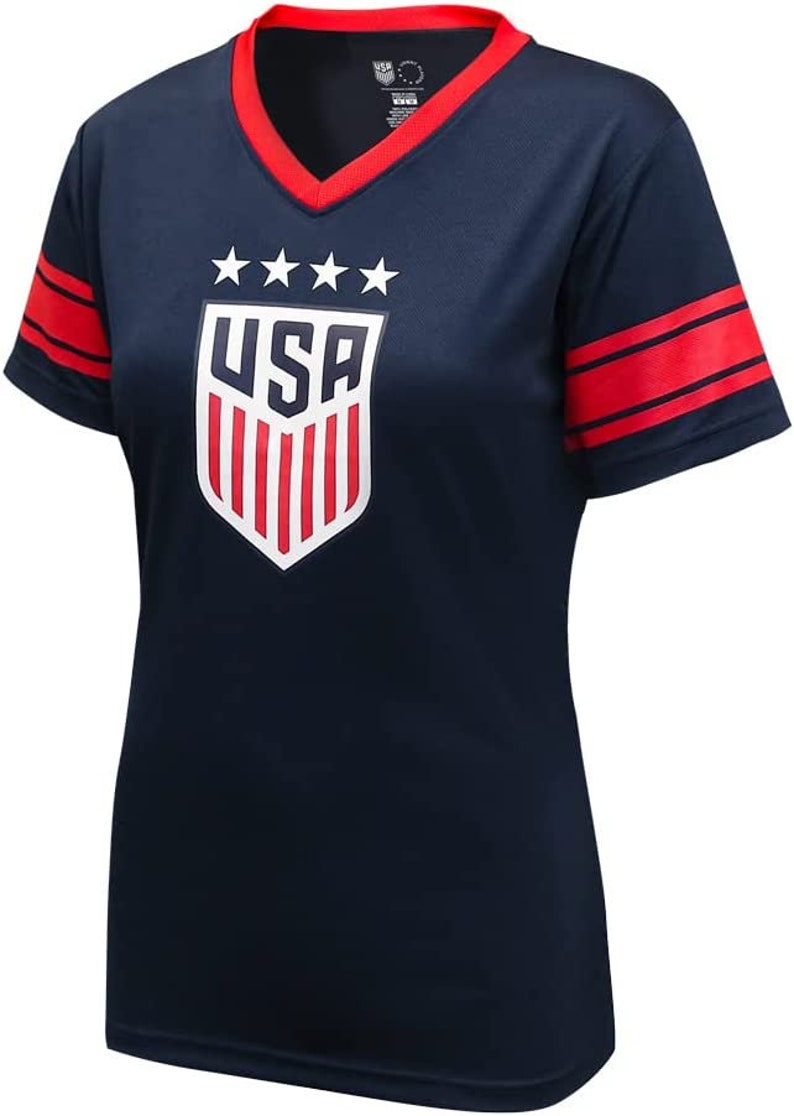 May include: A navy blue short-sleeved t-shirt with red and white accents. The shirt features a white shield with the word "USA" and five stars above it. The sleeves have two red stripes.