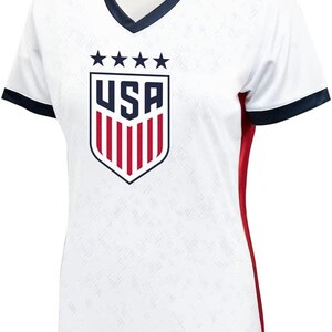 May include: White soccer jersey with a red, white, and blue USA crest and five stars above the crest. The jersey has red side panels and navy blue trim on the collar and sleeves.