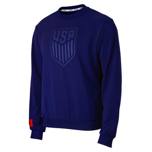 Op de afbeelding: Een marineblauw sweatshirt met een groot geborduurd USA-wapen op de voorkant. Het wapen heeft de vorm van een schild met de letters USA in het midden en verticale strepen aan de zijkanten. Het sweatshirt heeft een rode streep op de linkermouw.