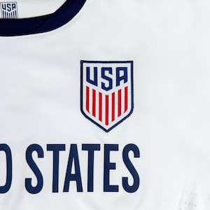 Pode incluir: Camisola de futebol branca com gola azul e as palavras "United States" impressas em azul abaixo de um escudo vermelho, branco e azul com as letras "USA" no seu interior.