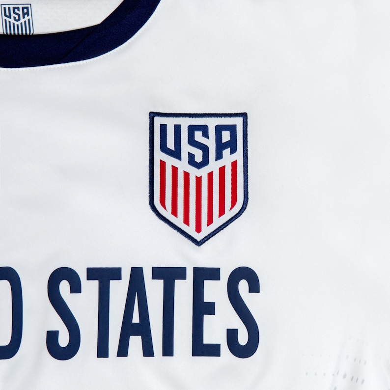 Pode incluir: Camisola de futebol branca com gola azul e as palavras "United States" impressas em azul abaixo de um escudo vermelho, branco e azul com as letras "USA" no seu interior.