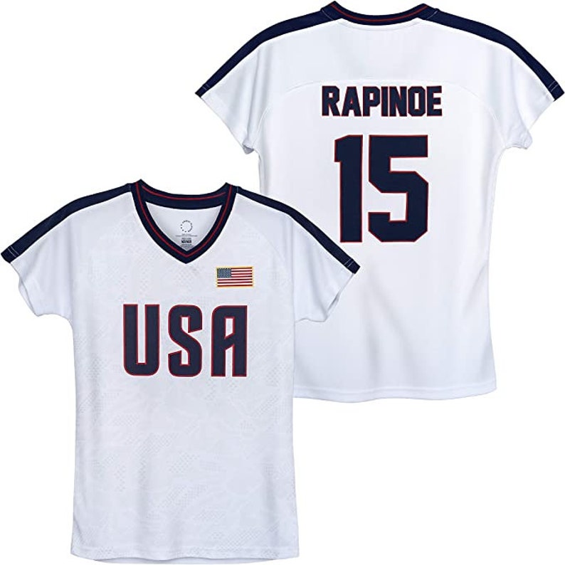 K&ouml;nnte beinhalten: Wei&szlig;es Fu&szlig;balltrikot mit blauen und roten Akzenten. Die Vorderseite des Trikots tr&auml;gt den Schriftzug "USA" in Blau mit einer kleinen amerikanischen Flagge. Die R&uuml;ckseite des Trikots tr&auml;gt den Namen "RAPINOE" und die Nummer "15" in Blau.