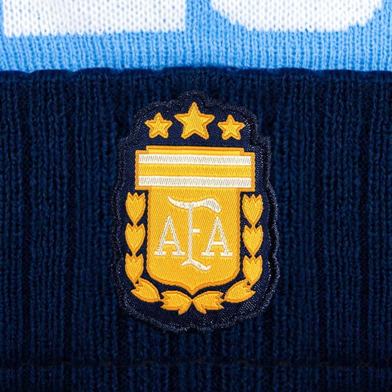 Puede incluir: Un gorro de punto azul marino con una banda azul claro y blanca. El gorro presenta un parche con el emblema de la Asociaci&oacute;n del F&uacute;tbol Argentino, que incluye estrellas doradas, un escudo y las letras "AFA".