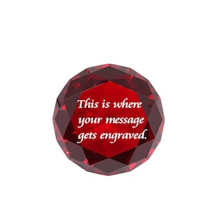 May include: A faceted, ruby-red crystal paperweight with the text "This is where your message gets engraved." The crystal is round and has many reflective surfaces. The background is plain white.