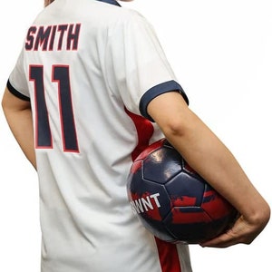 May include: A white soccer jersey with red and blue accents. The jersey has the name "SMITH" and the number "11" printed on the back. A person is holding a soccer ball in their right hand.