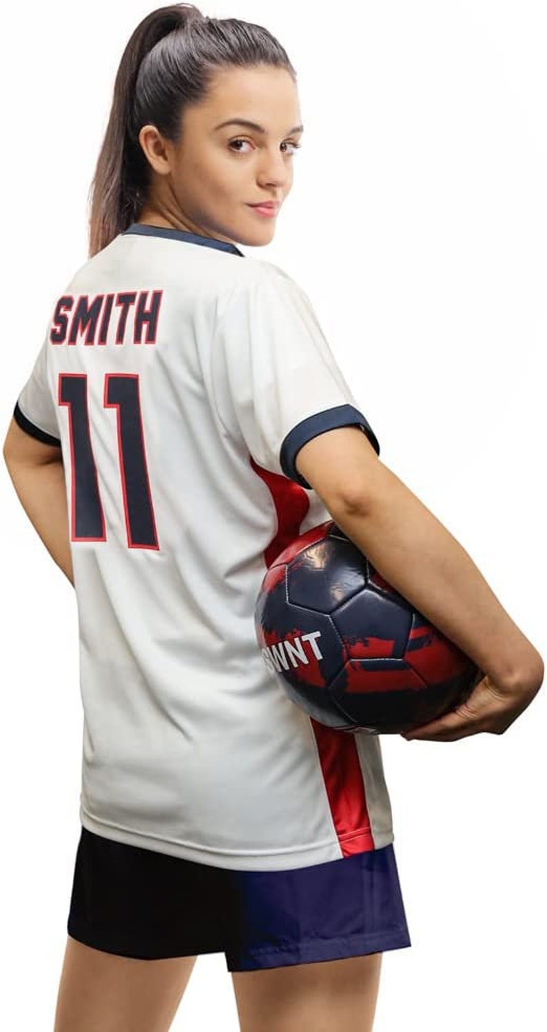 May include: A white soccer jersey with red and blue accents. The jersey has the name "SMITH" and the number "11" printed on the back. A person is holding a soccer ball in their right hand.